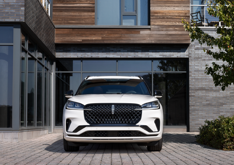 A white 2025 Lincoln Aviator® SUV is shown parked outside a modern building with gray brick and wood accents. | Doggett Lincoln of Beaumont in Beaumont TX